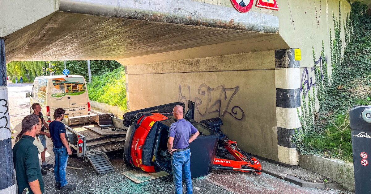 Graafmachine valt van aanhanger na botsing met brug